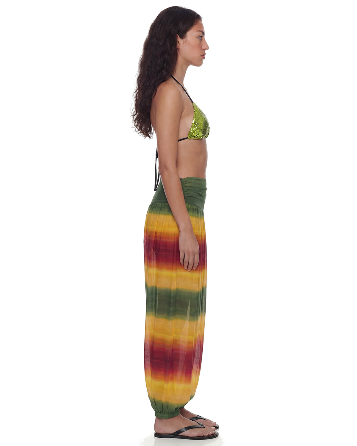 Modelo de perfil com a Calça Aladdin Tule Ginga em degradê verde, amarelo e vermelho, mostrando o caimento largo e a cintura franzida