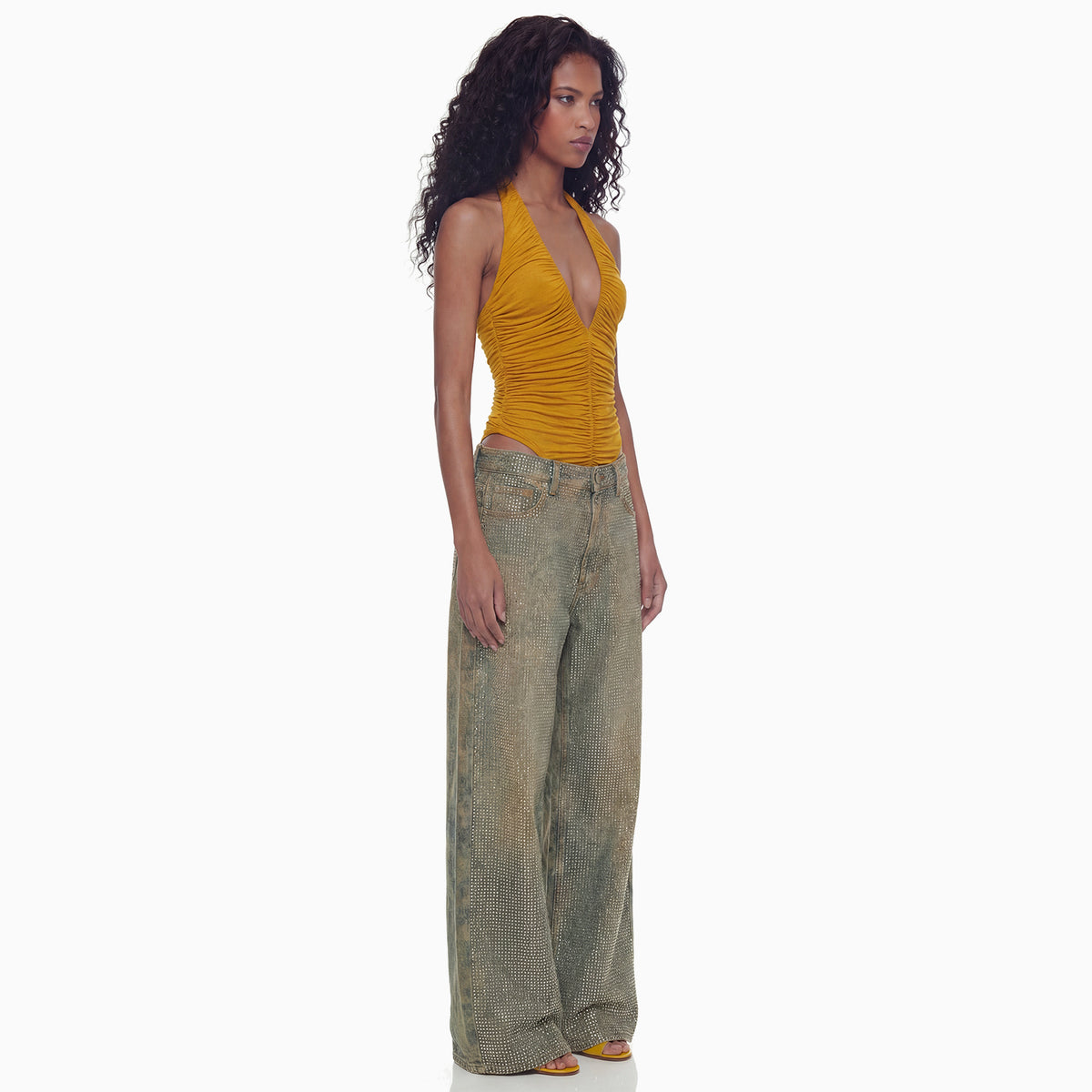 Modelo de perfil com a Calça Jeans Strass pantalona brilhante e body halter mostarda de decote em V