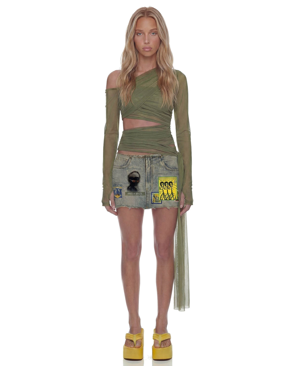 Modelo de frente veste a Mini Saia Jeans Brasilidades de cintura baixa, jeans claro com patches, barra desfiada e bem curta