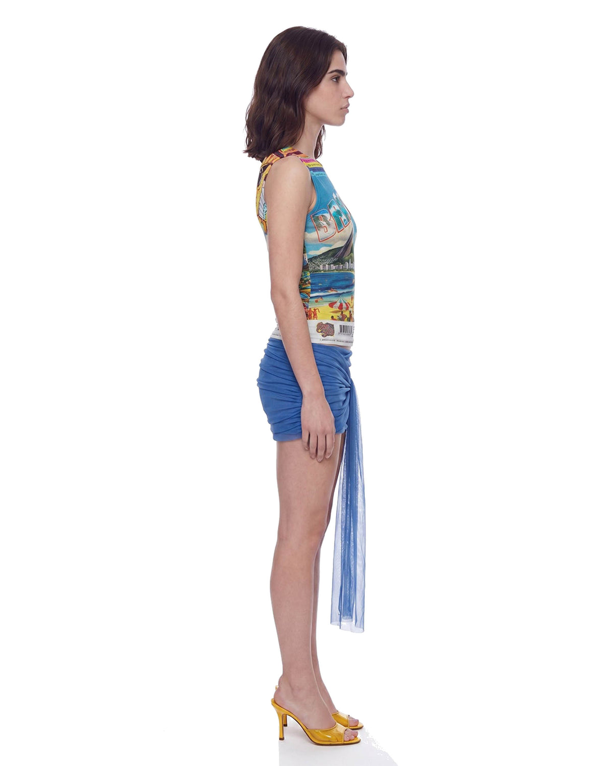 Vista lateral de perfil da Mini Saia Transpasses Tule Hexa azul na modelo, comprimento curto e faixa de tule esvoaçante