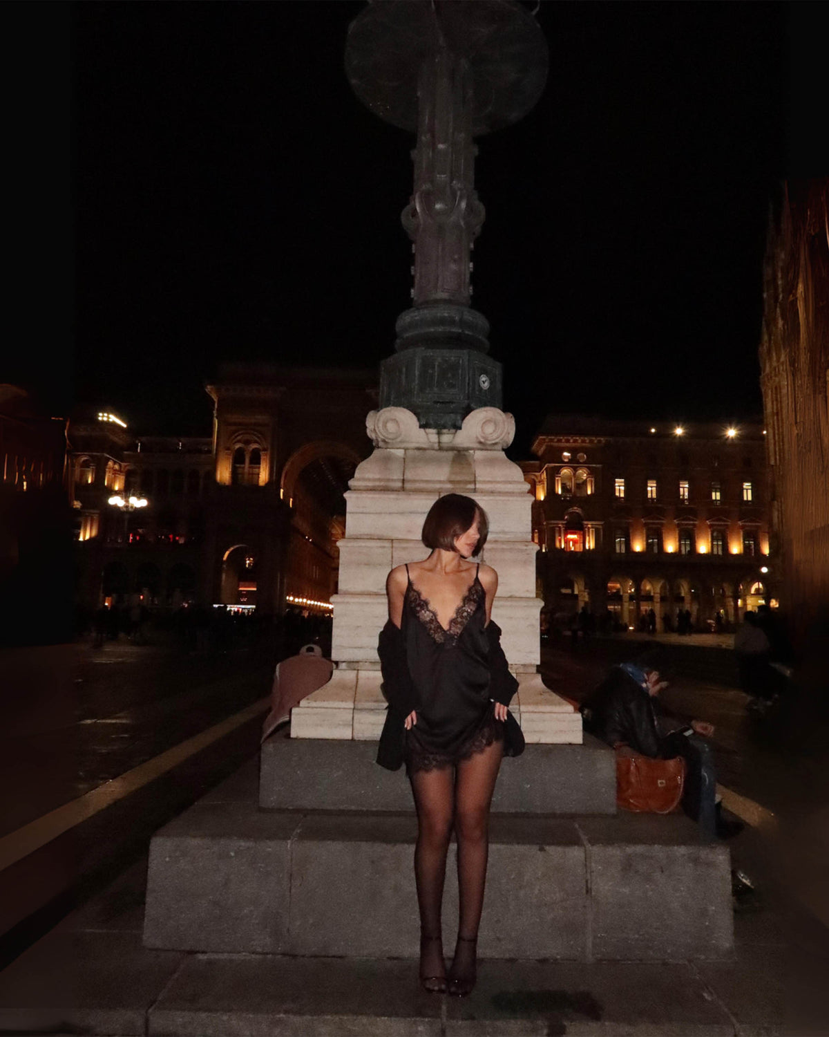 Modelo posa com o Vestido Affair preto acetinado curto e casaco sobre os ombros, decote em renda