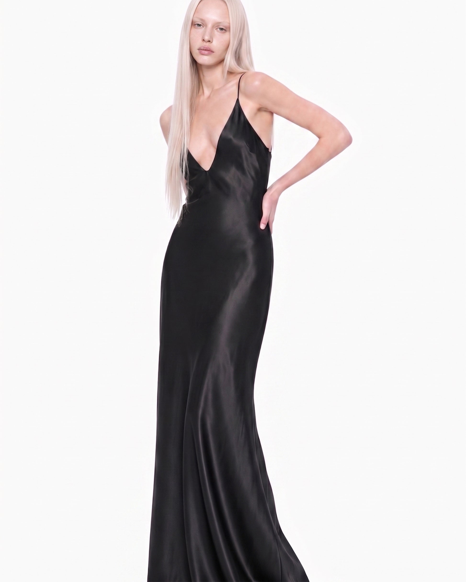 Modelo em três quartos com o Vestido Ivo preto, mão na cintura, decote V profundo e caimento acetinado